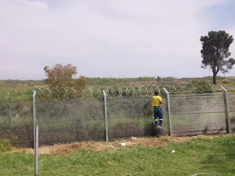 Worker installing razor wire coils on top of a galvanised security fence at an industrial site in Cape Town